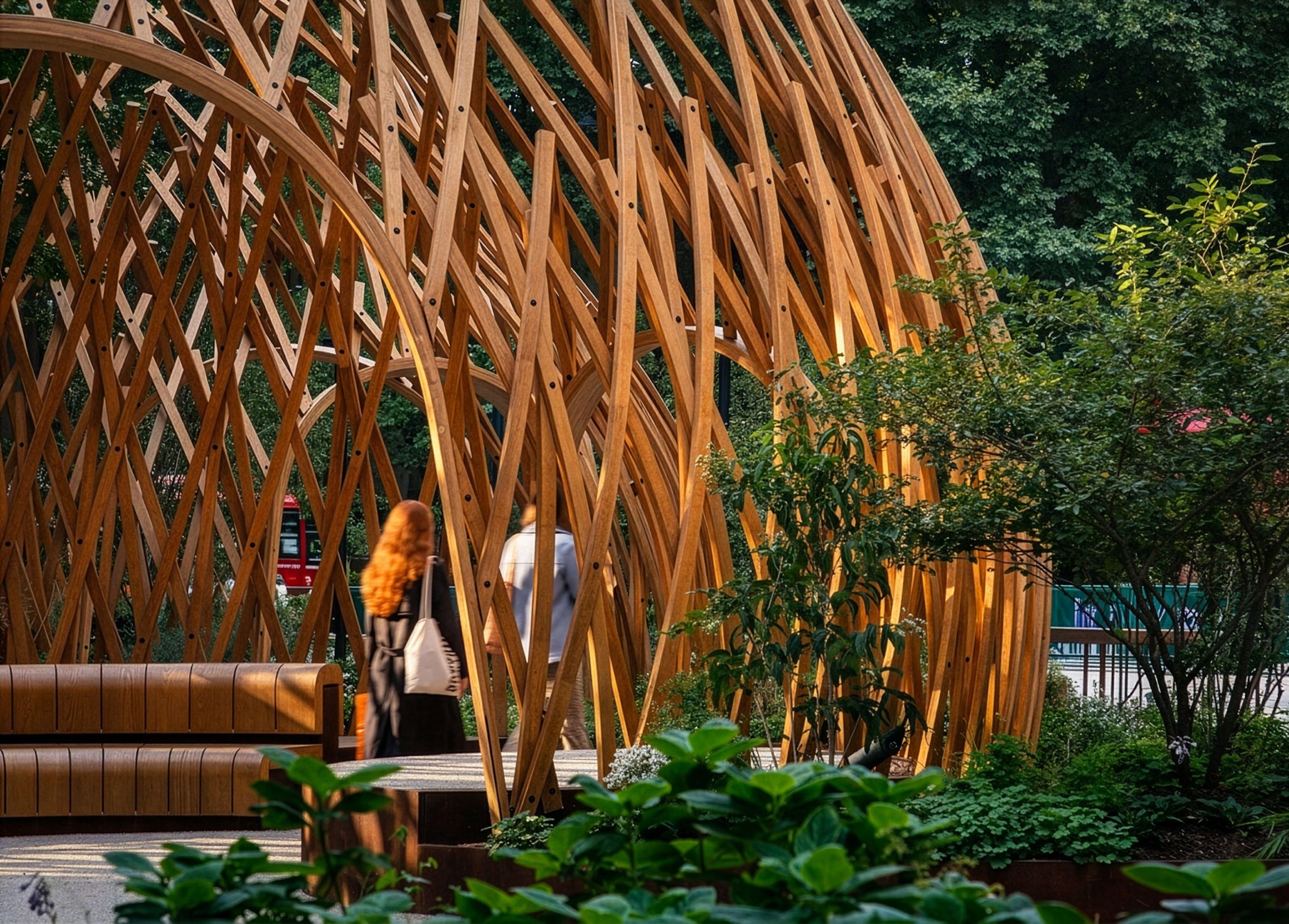 Curved timber members of a lattice canopy meeting the ground beside dense planting and timber seating, two people passing through
