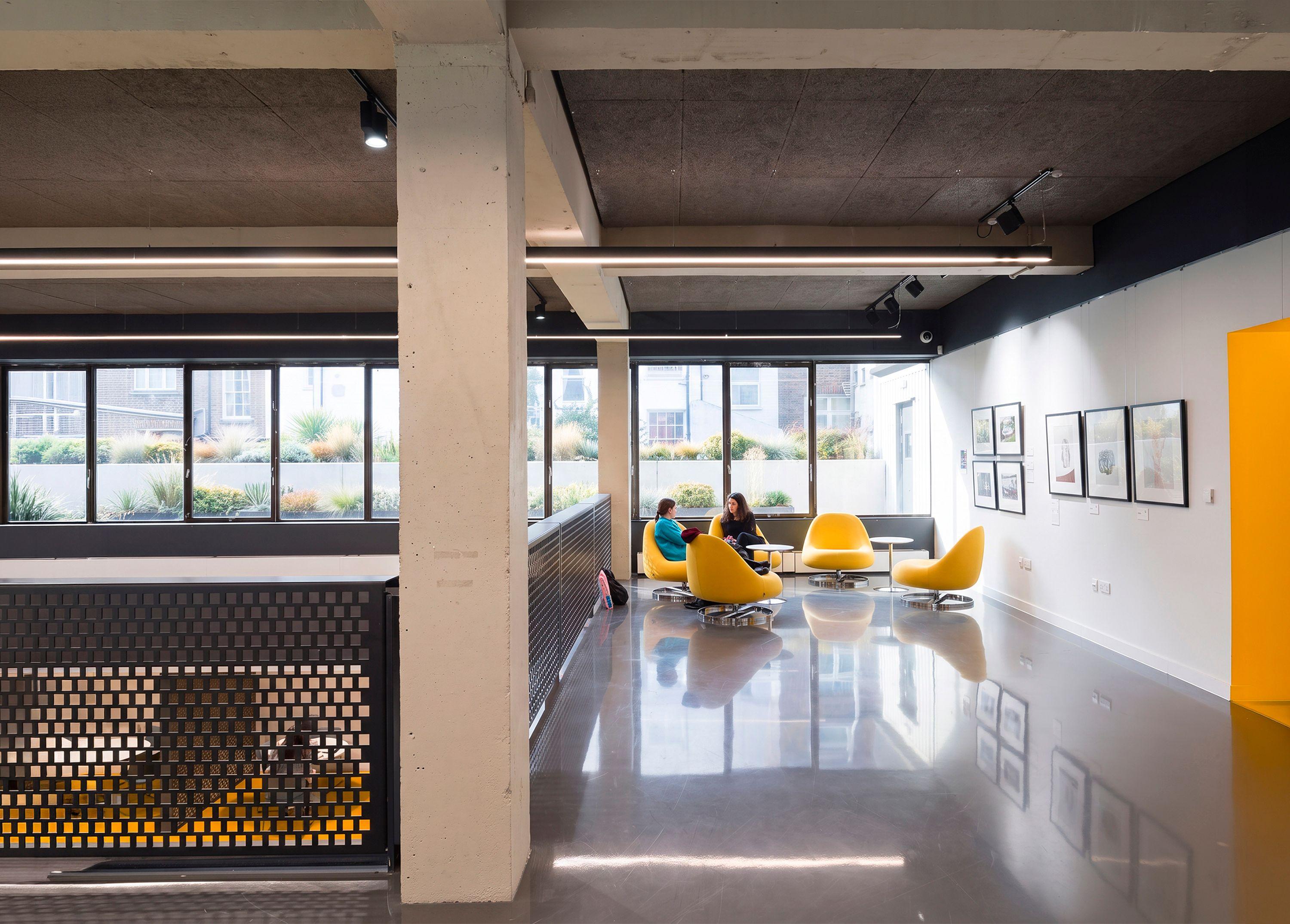 Gallery area in Brighton reflected in the dark polished resin floor, with yellow chairs, a perforated balustrade over the double-height opening and a dark woodwool ceiling above.